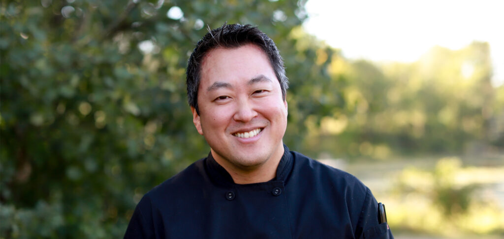 Chef Ryan Schmitt, director of culinary services at Elim Shores in Eden Prairie, MN, smiles warmly outdoors in his black chef’s jacket, surrounded by natural greenery.
